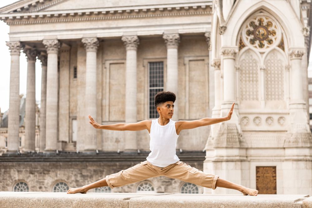 Dancer performs a graceful split leap in front of ornate classical architecture with stone columns and decorative details.