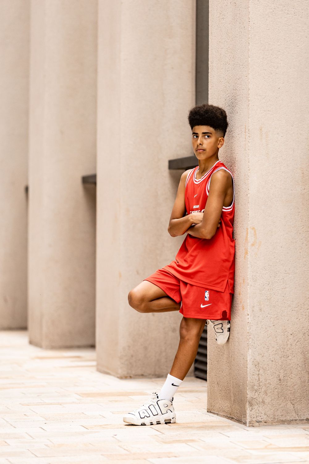 Athletic figure in red basketball uniform leans against concrete pillar in urban setting wearing white sneakers.