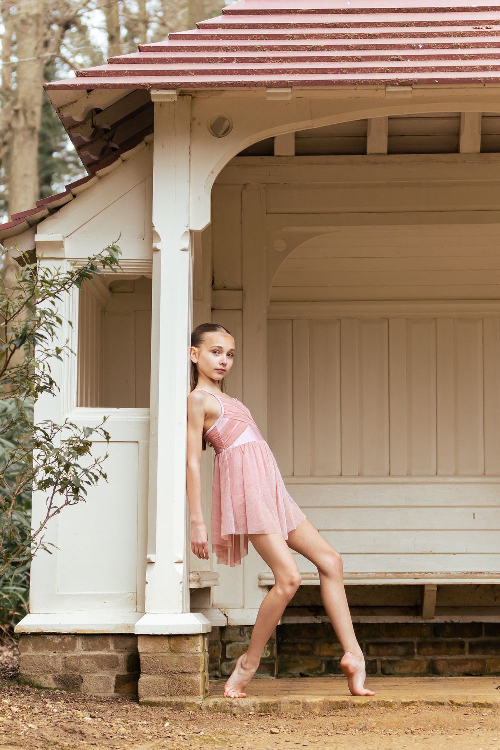 A ballet dancer in a pink dress poses gracefully on the steps of a vintage gazebo with white pillars and striped roof.