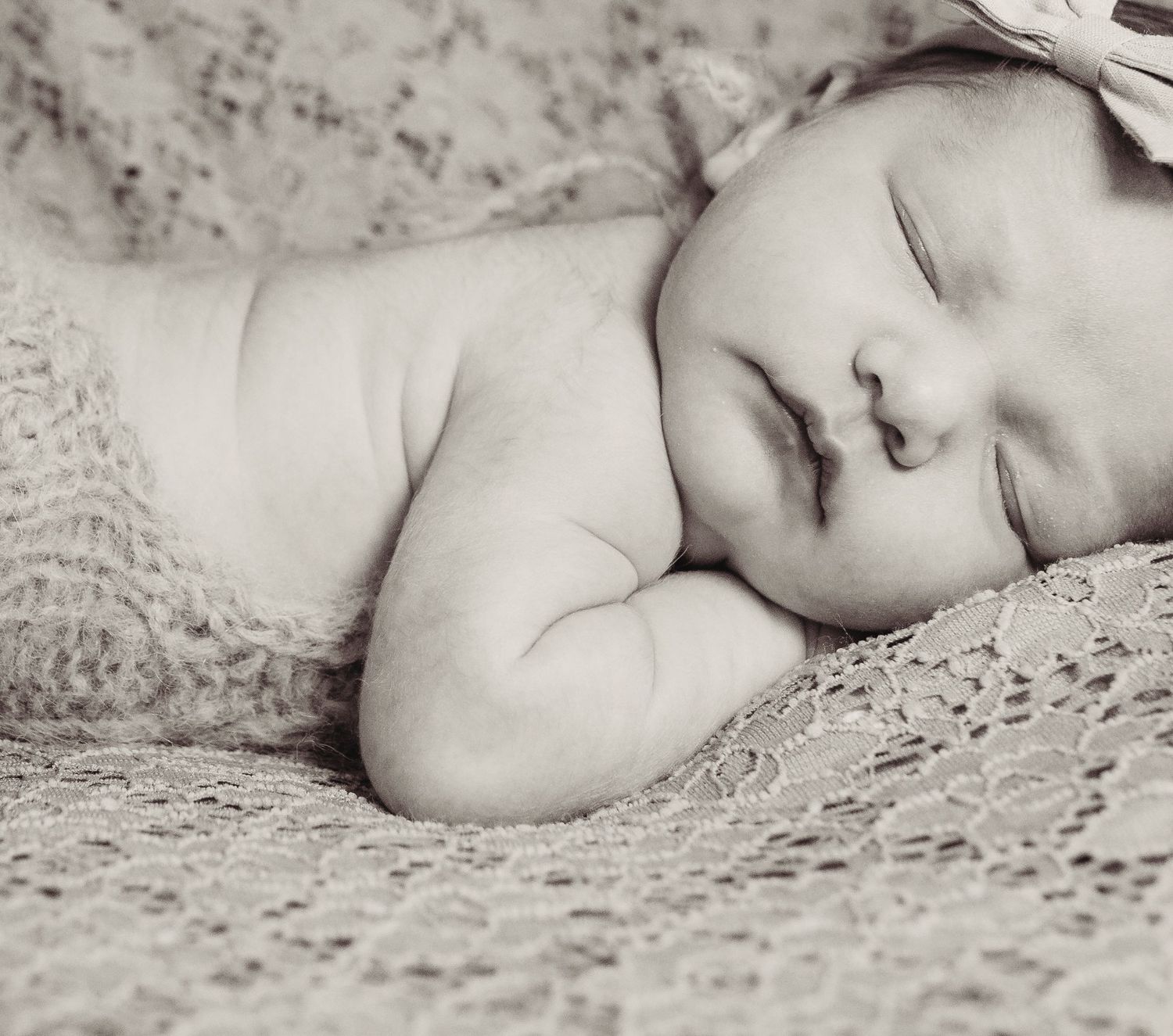 A sleeping baby rests peacefully on lace fabric in a gentle black and white portrait.
