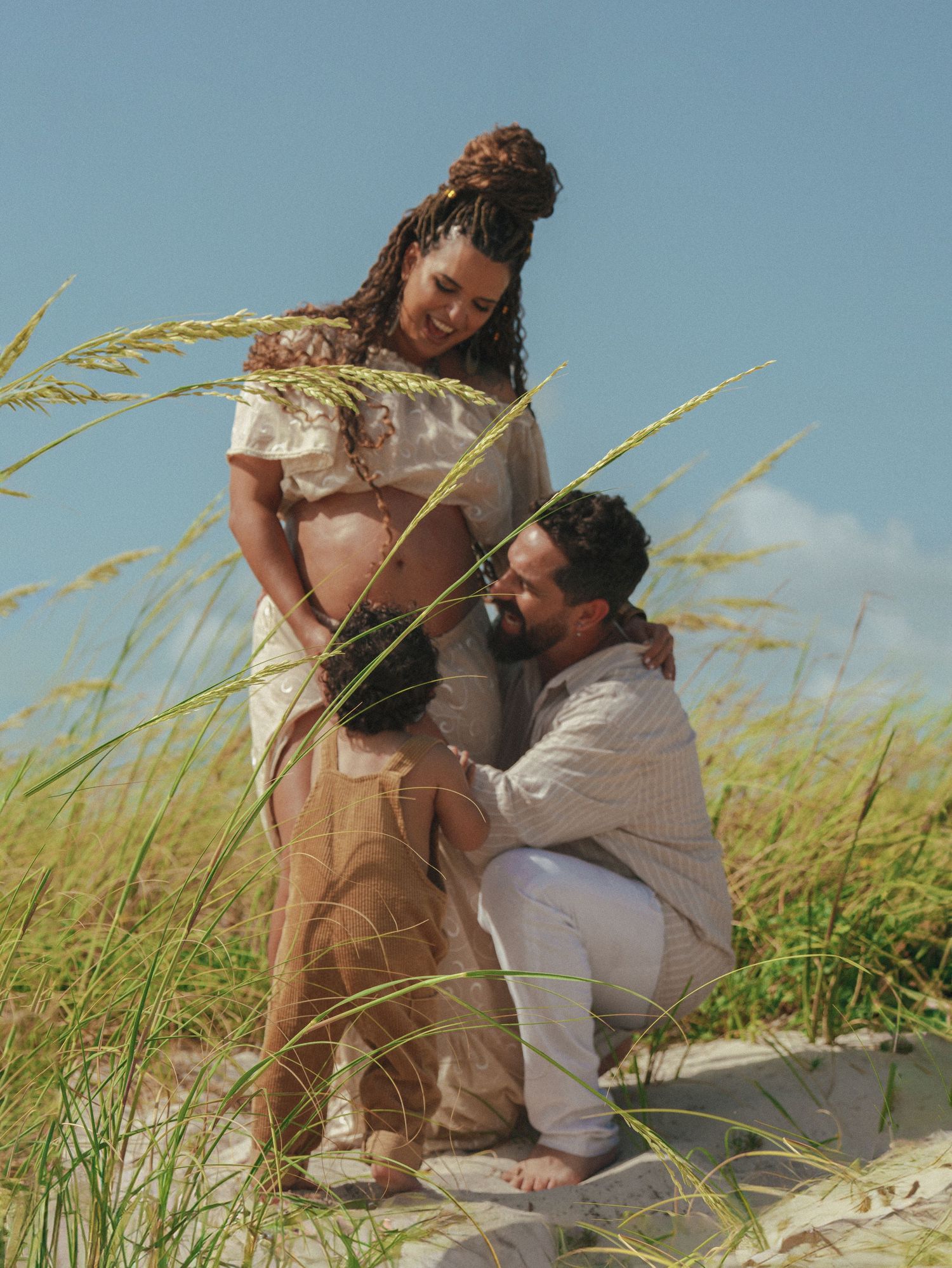 A tender maternity moment captured in tall grass against a blue sky background.