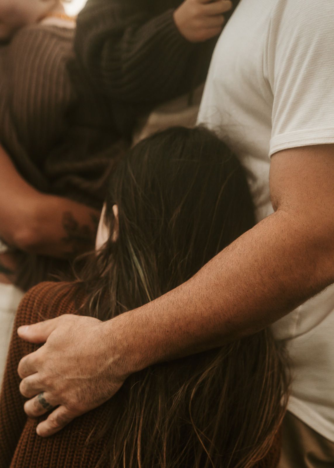 Person in a white t-shirt comforting someone by placing a gentle hand on their long dark hair at golden hour