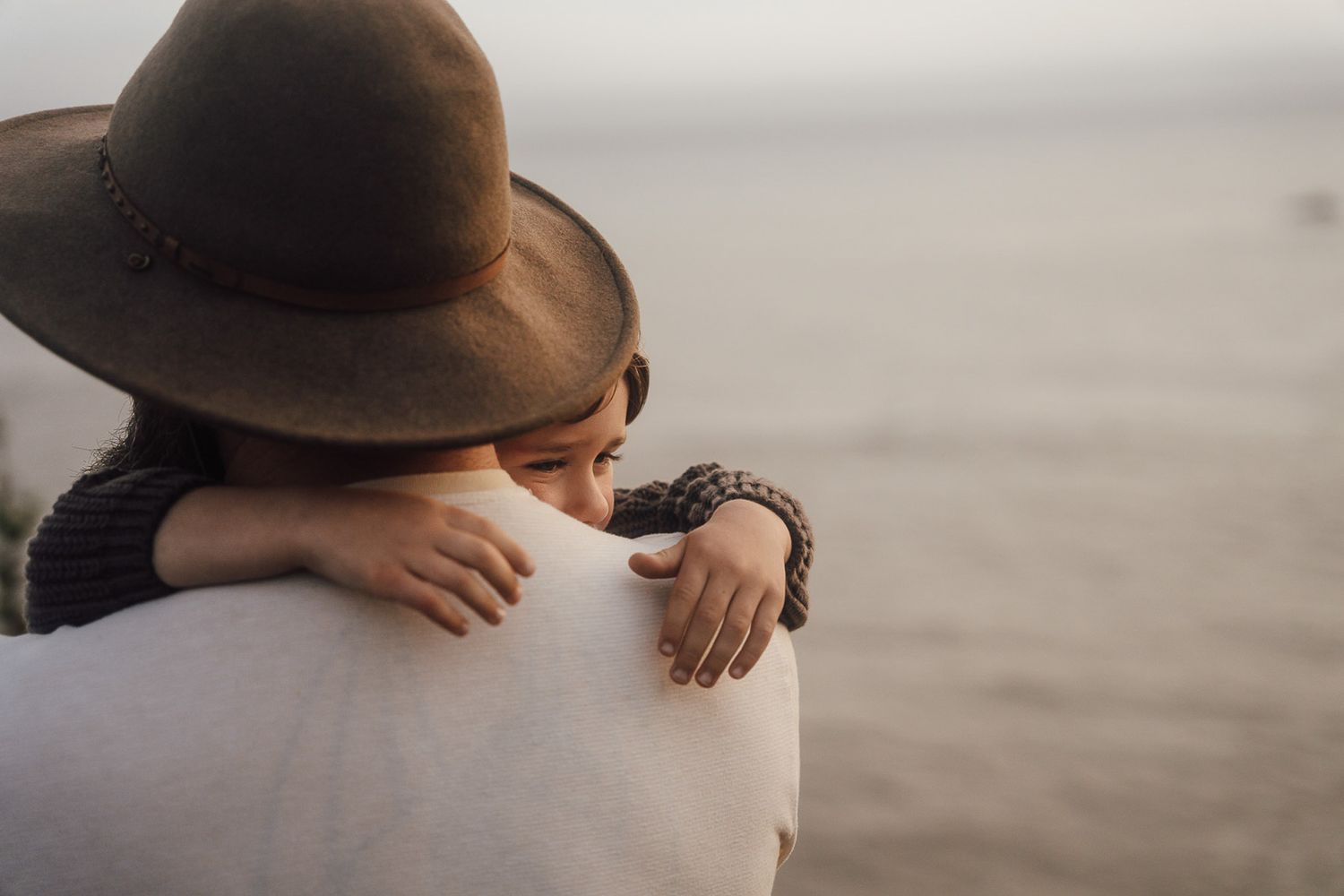 A close embrace captured from behind, showing a person wearing a brown hat at golden hour by the water.