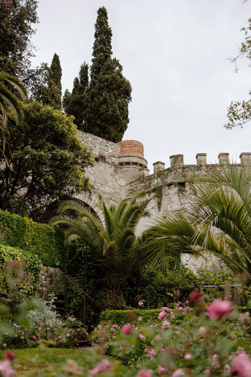 Medieval castle walls surrounded by lush palm trees and blooming pink flowers in a Mediterranean garden setting.