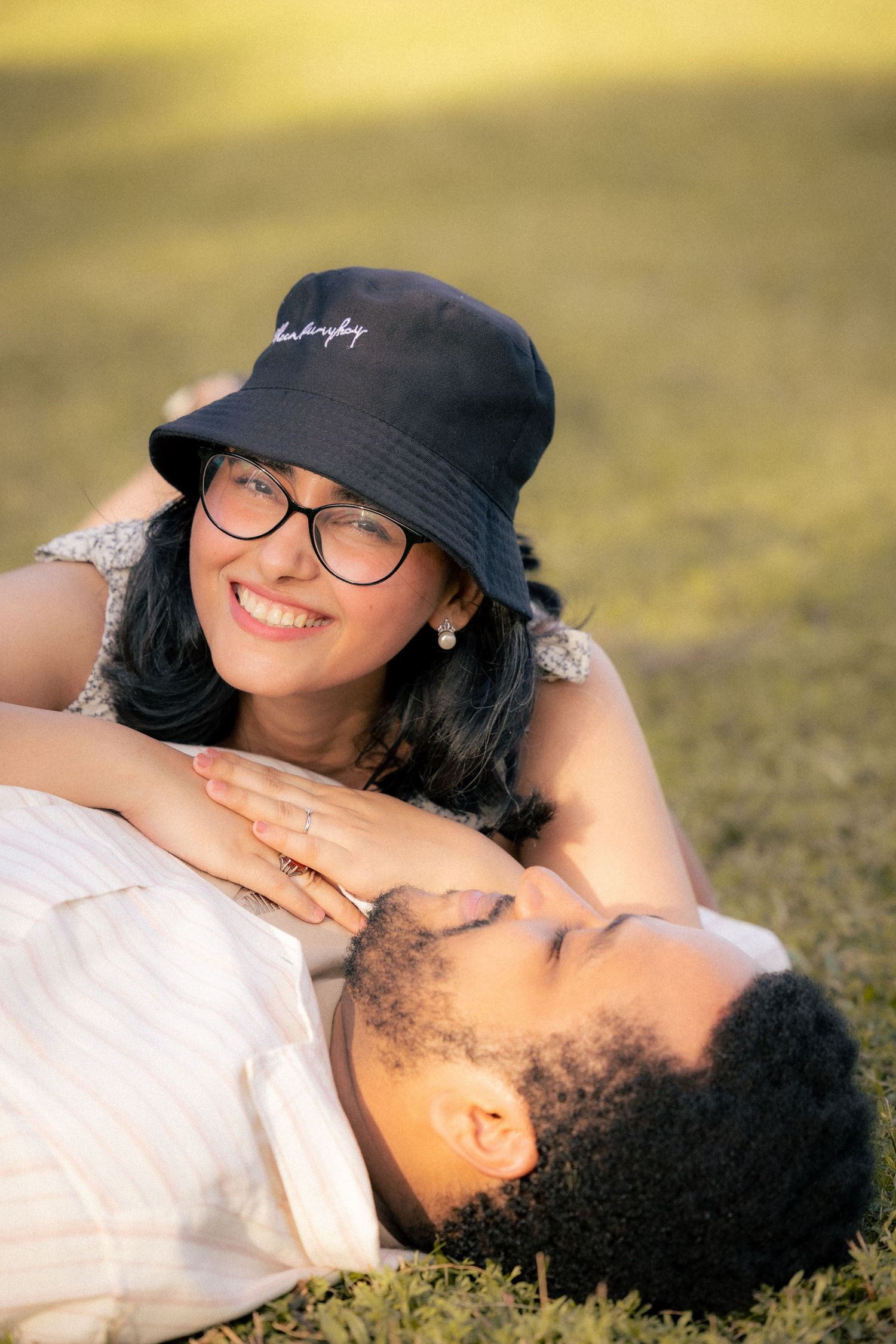 Two people lying on grass outdoors on a sunny day, one wearing a black bucket hat and glasses.