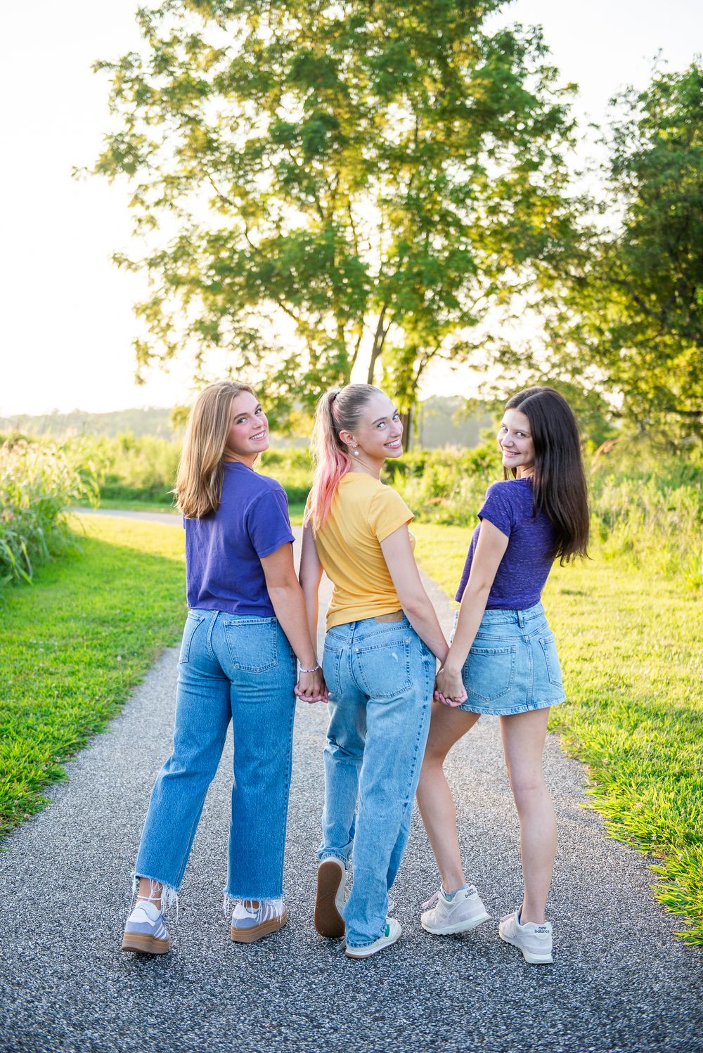 Three friends in denim showing their backsides while standing together on a gravel path during golden hour lighting.