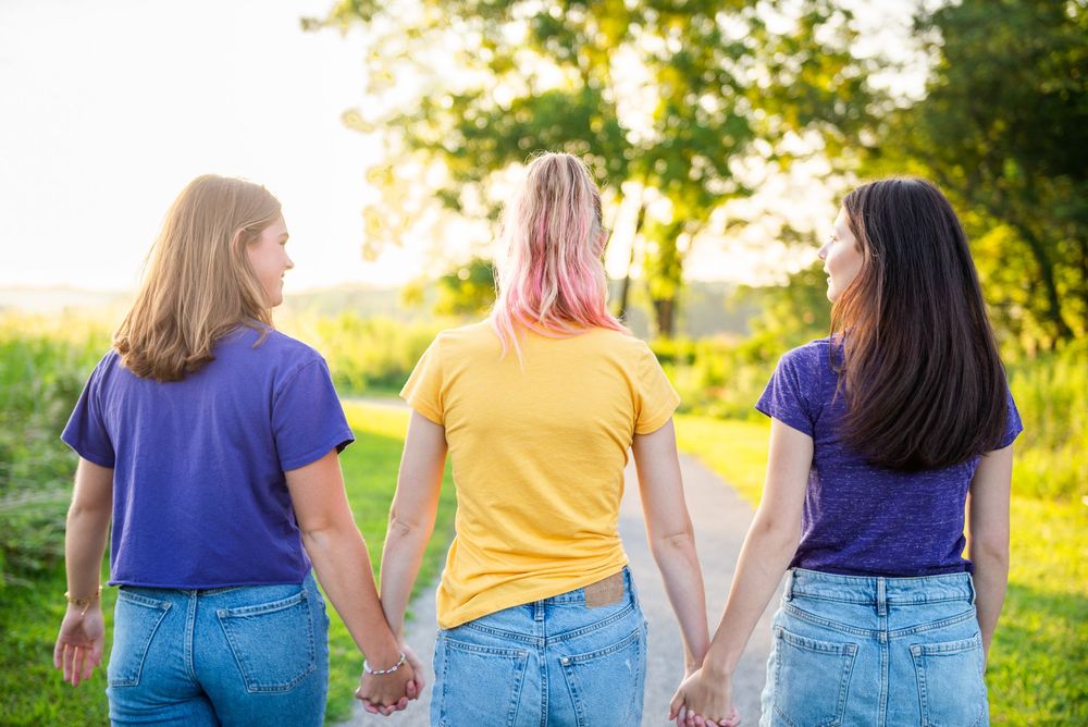 Three women walking away hand in hand while wearing blue jeans and t-shirts in a sunny outdoor setting.