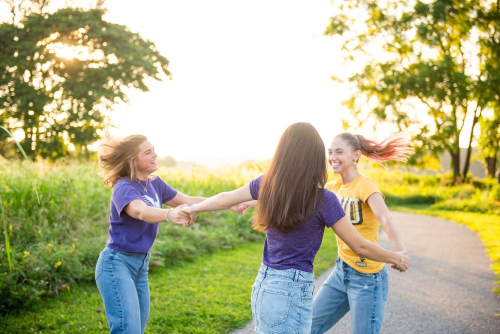 Three friends joyfully dancing and holding hands together on a nature path at sunset.