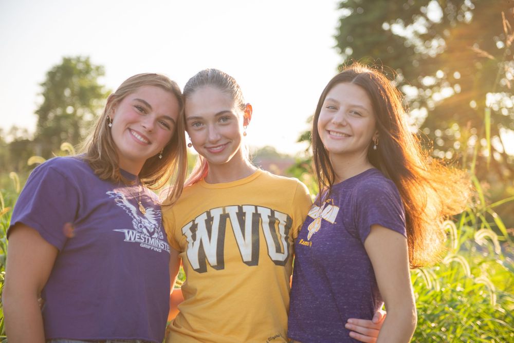 Three students in navy blue and yellow college t-shirts smile together outdoors during golden hour sunset.