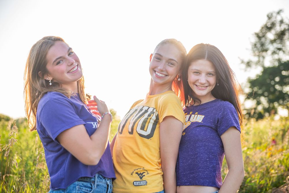 Three friends pose together in casual outfits against a sunlit summer meadow background.