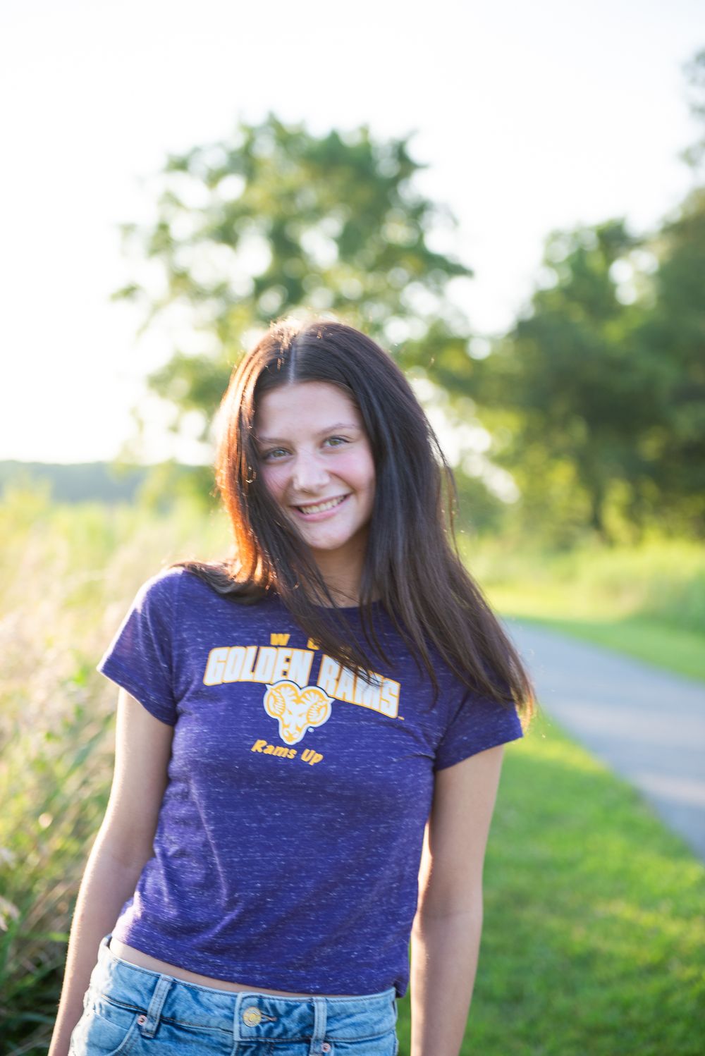 A person in a navy blue graphic t-shirt smiles while standing on a path with sunlit greenery in the background.