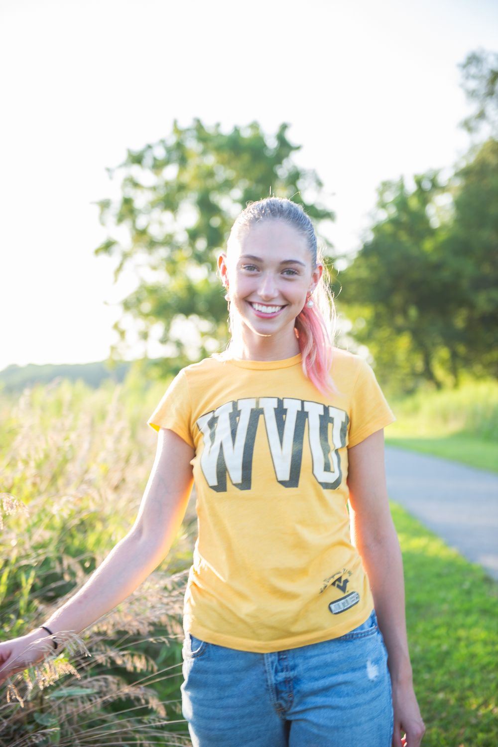 Person wearing a yellow WVU t-shirt and blue jeans stands smiling on a path with greenery in the background.