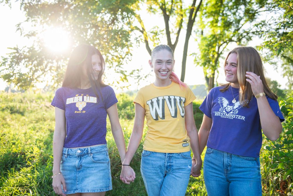 Three friends holding hands and laughing together in a sunny park while wearing matching jeans and colorful t-shirts.