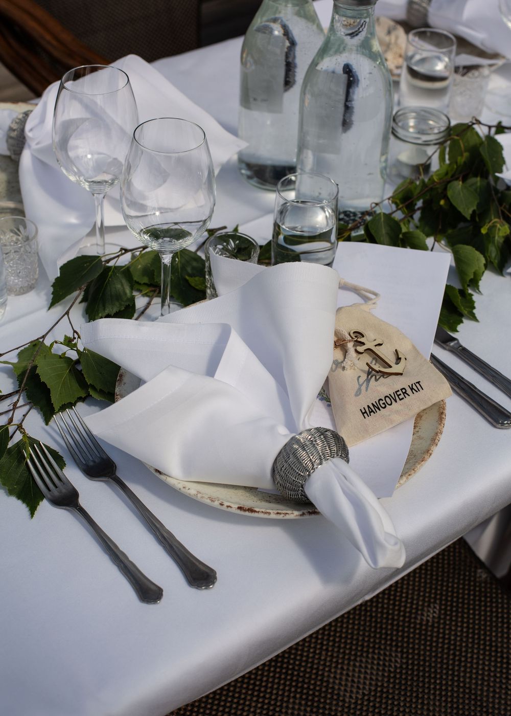 Elegant white wedding table setting with folded napkin, greenery garland, and hangover kit favor.