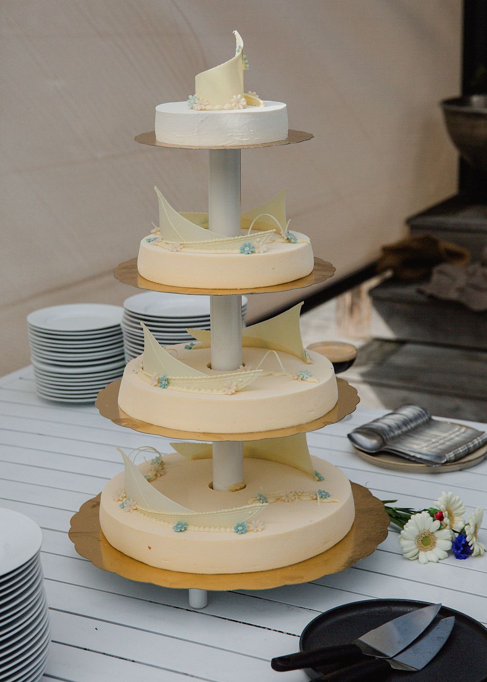 Five-tier white wedding cake with decorative ribbon accents displayed on white table outdoors.