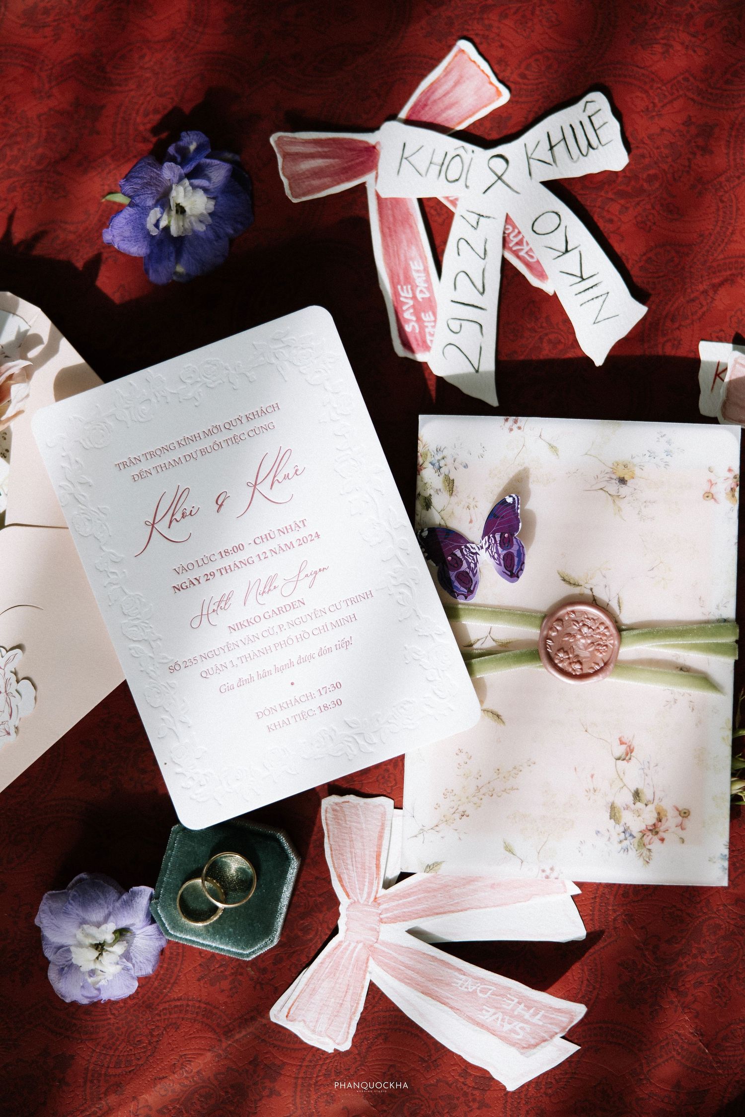 Elegant wedding invitation suite with floral details, wax seal, pink ribbons, and gold rings on red fabric.