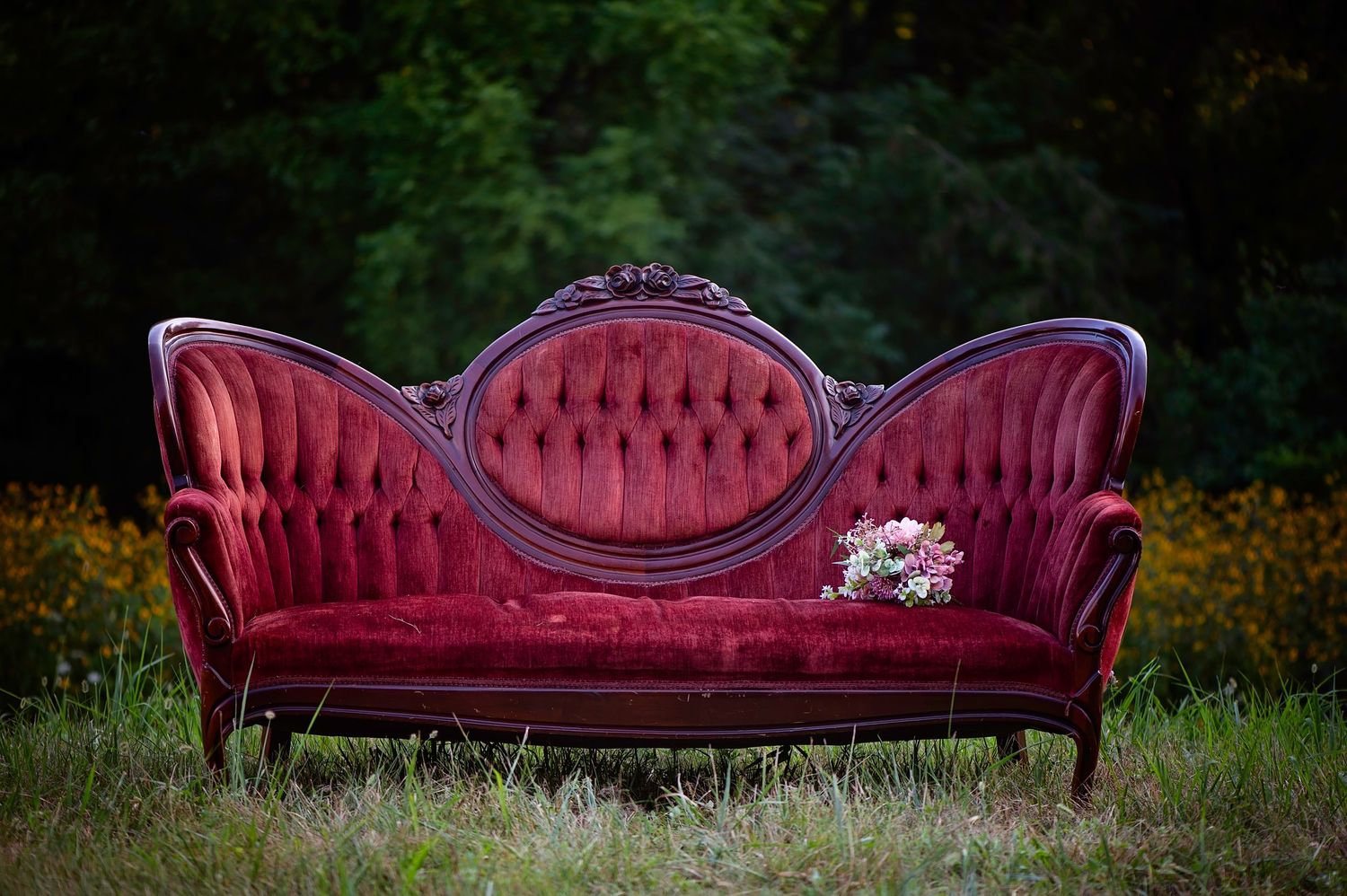 A vintage red velvet Victorian sofa with ornate wooden frame sits alone on grass with dark foliage background.
