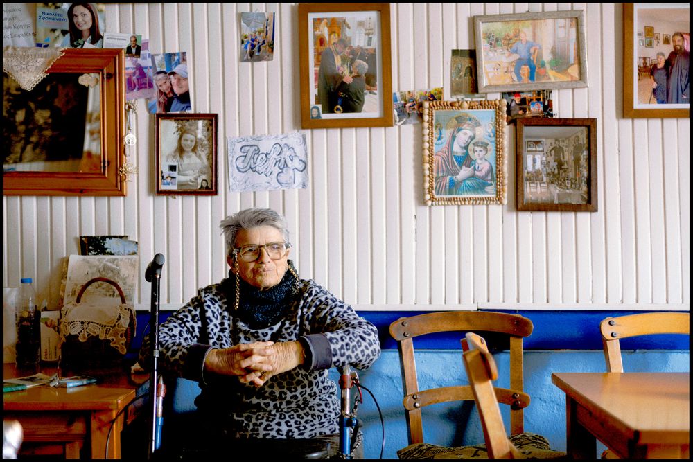 Elderly woman sitting inside a traditional Greek home surrounded by family photographs and religious icons.