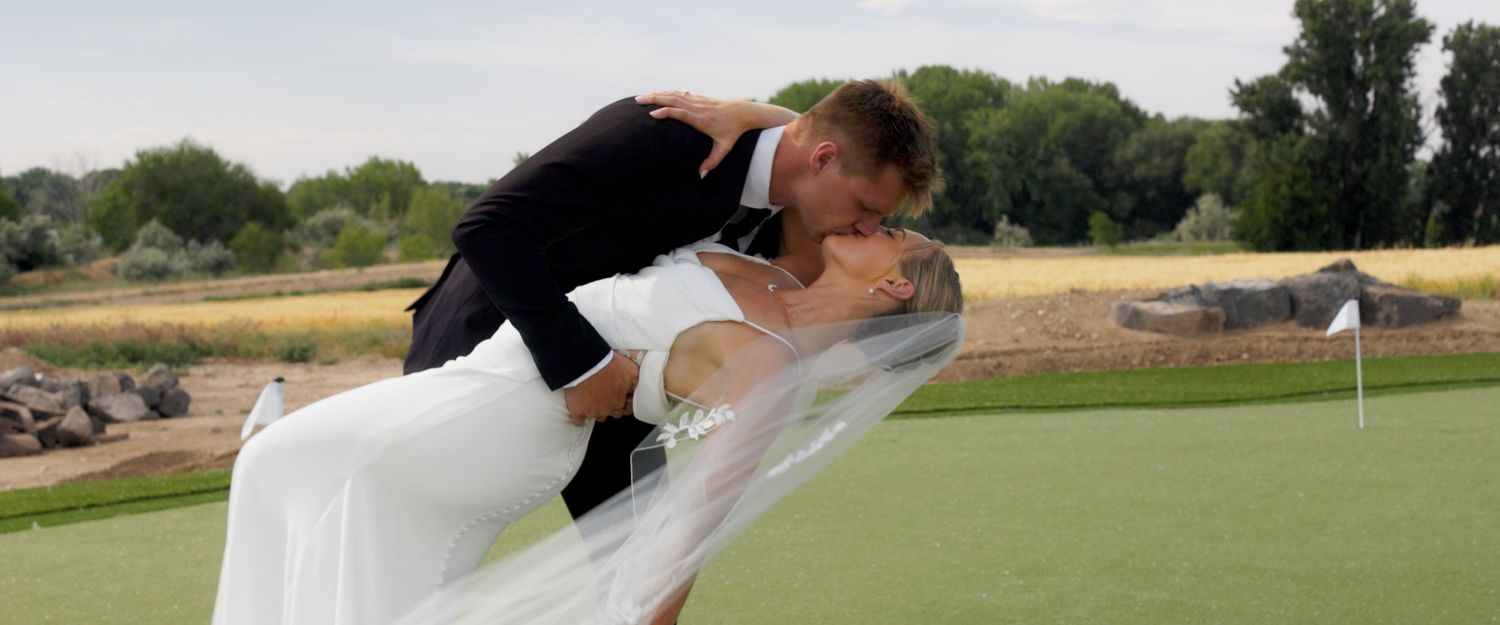 A romantic wedding couple shares a kiss while dipping on a scenic golf course with green grass and trees in the background.