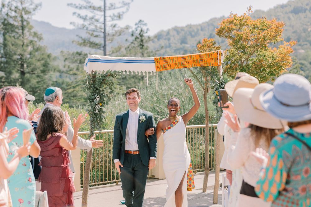 Just married couple walks under a colorful canopy as guests cheer, with Geneva Terrace’s mountain backdrop behind them.