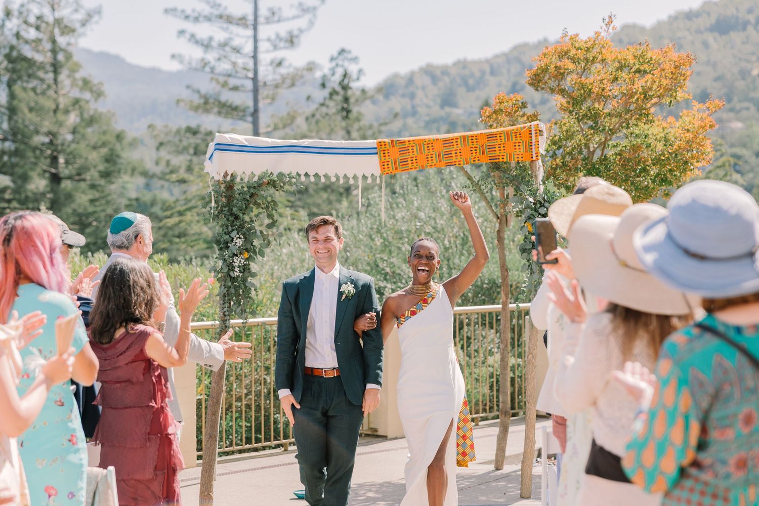 Just married couple walks under a colorful canopy as guests cheer, with Geneva Terrace’s mountain backdrop behind them.