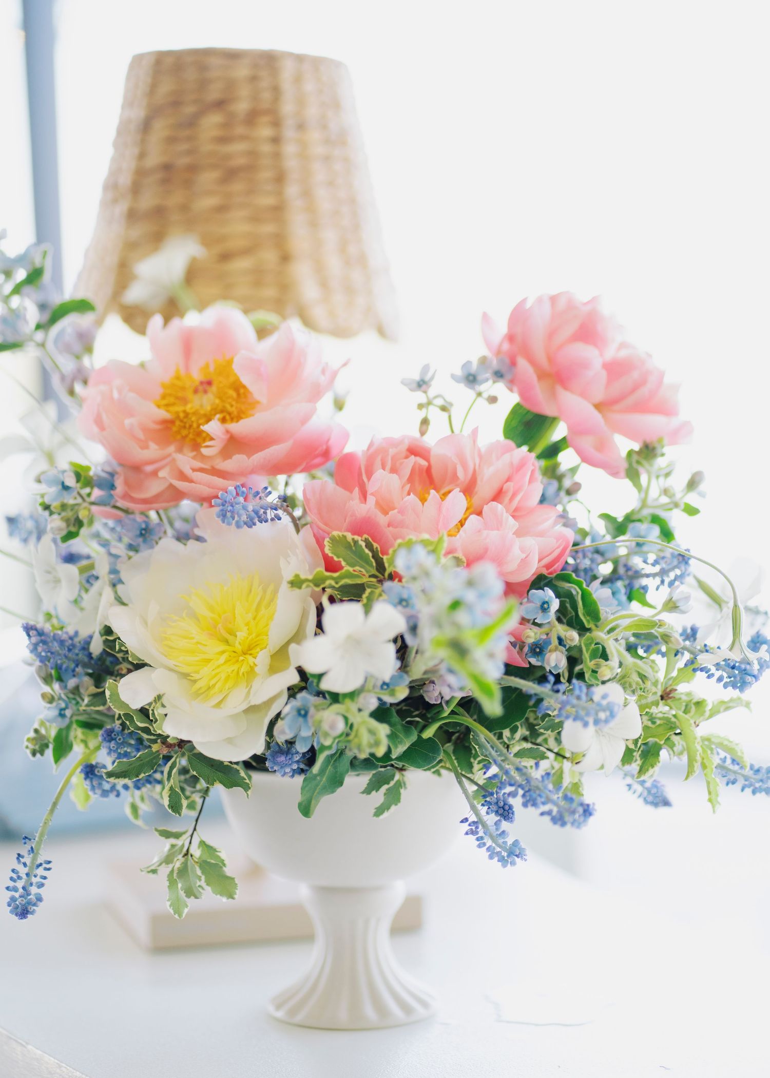 A elegant white pedestal vase holds pink peonies, yellow flowers, and blue forget-me-nots.