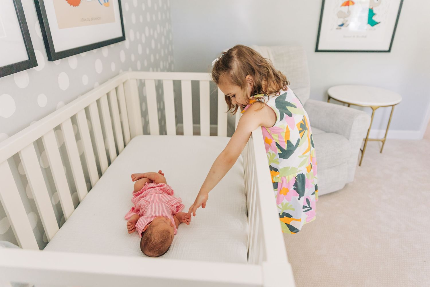 A toddler in a colorful dress reaches into a white crib where a baby is lying.
