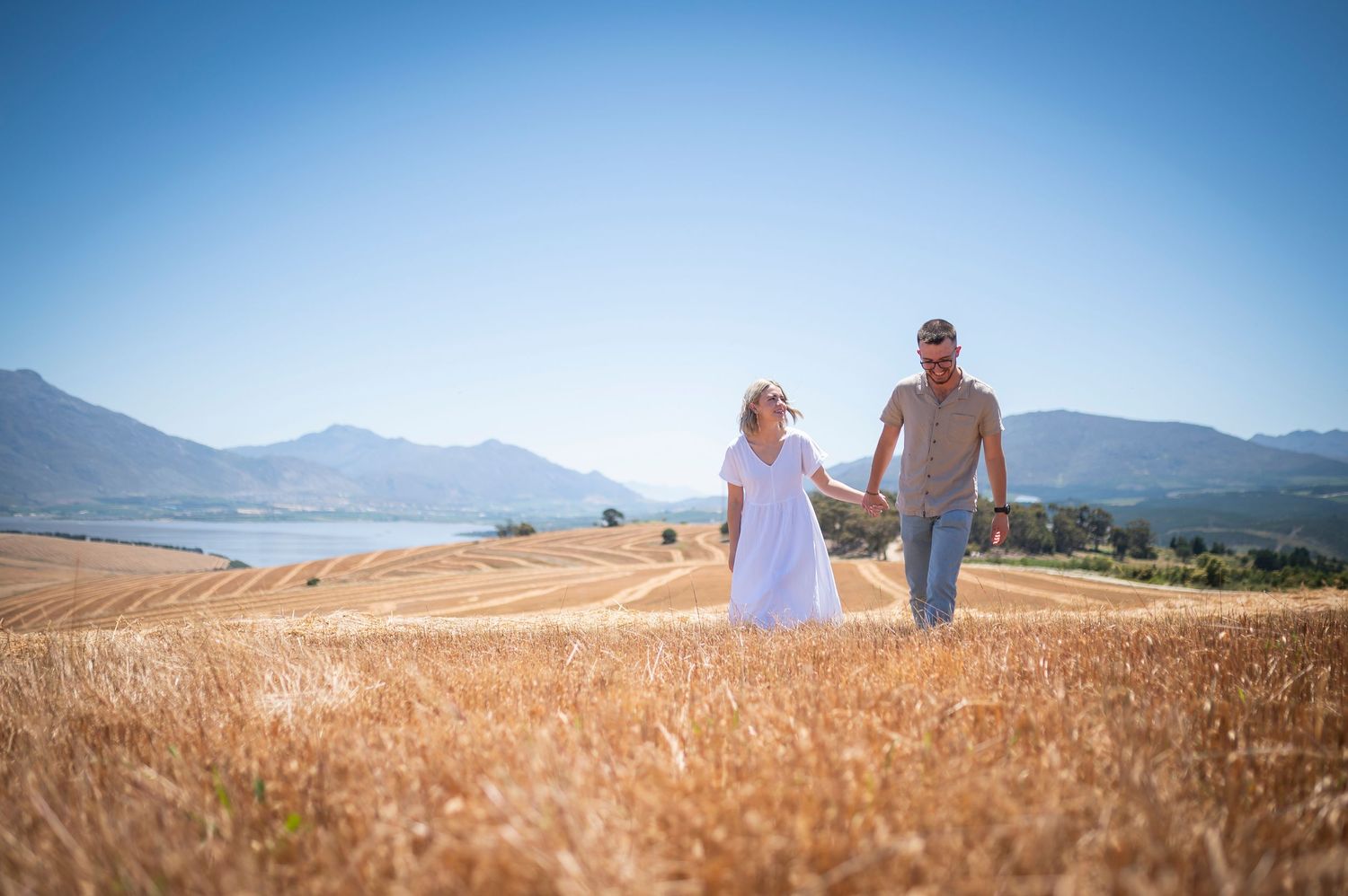 In a golden wheat field with mountain backdrop, two people walking under bright summer sky.
