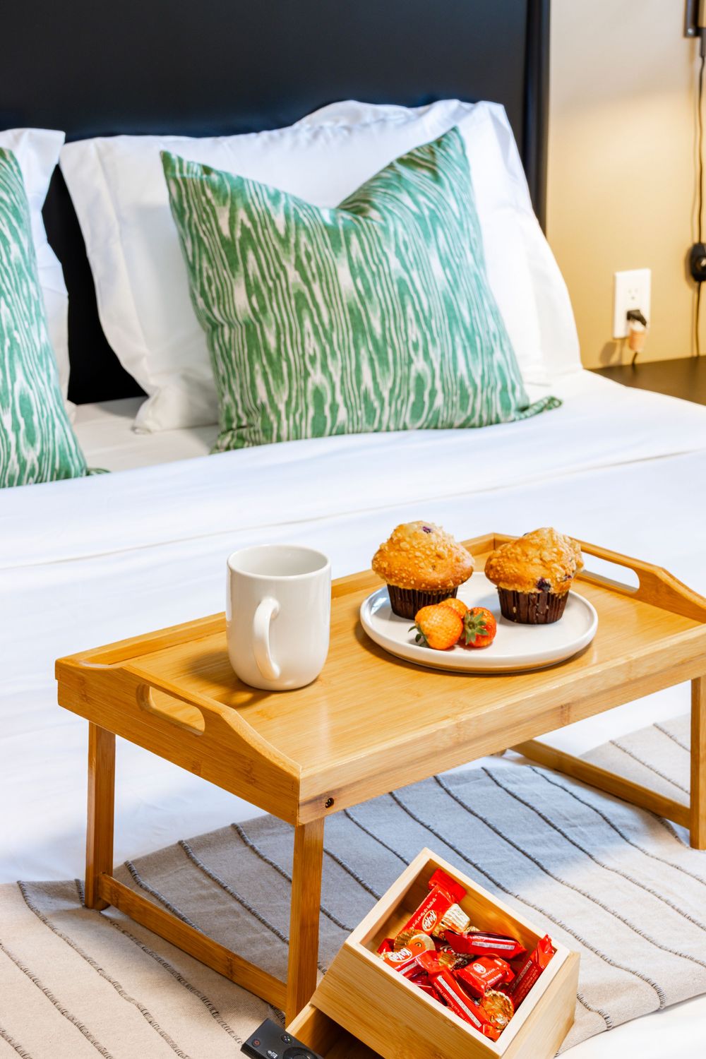 Wooden breakfast tray with muffins and coffee cups on white bedding with green tropical print pillows.