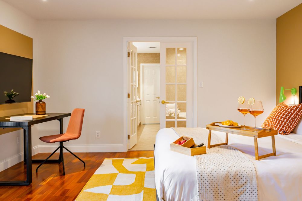 Bright bedroom interior with white walls, wooden floors, and natural light streaming through windows.