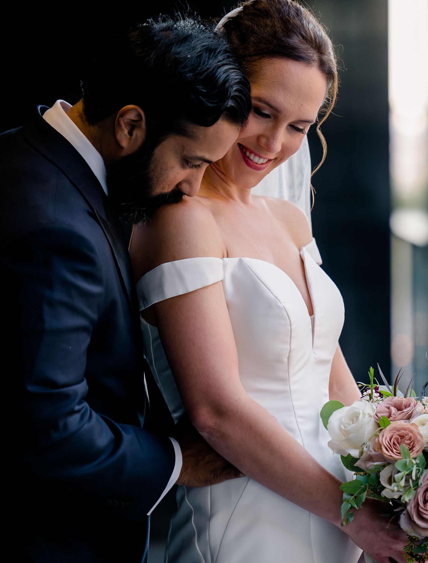 A tender moment between newlyweds as they embrace, with the bride wearing an off-shoulder white gown and holding a pink bouquet.