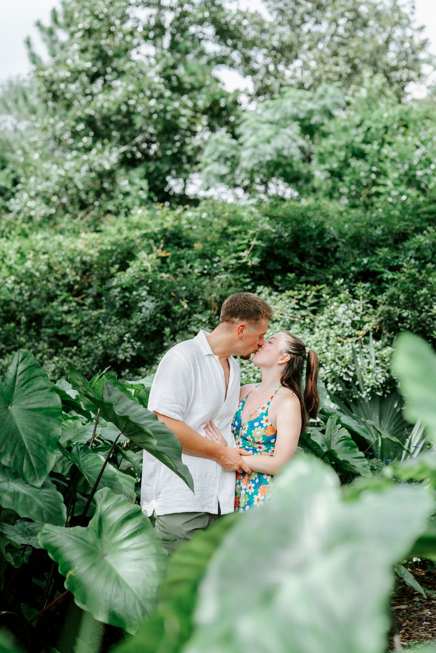 A romantic couple shares a tender moment among lush greenery in a garden setting.