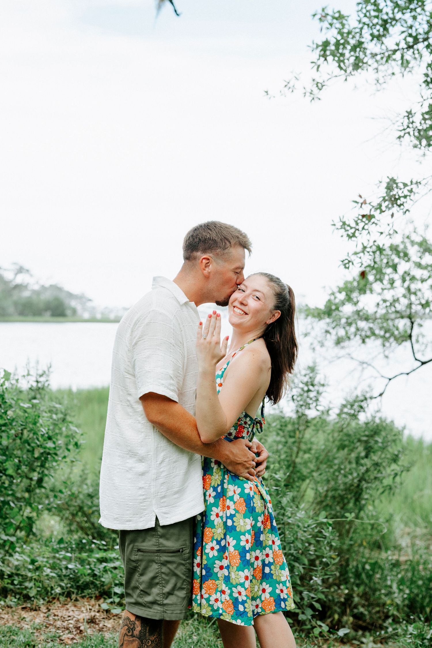 A joyful couple laughs together in a sunny outdoor setting with trees and flowers.