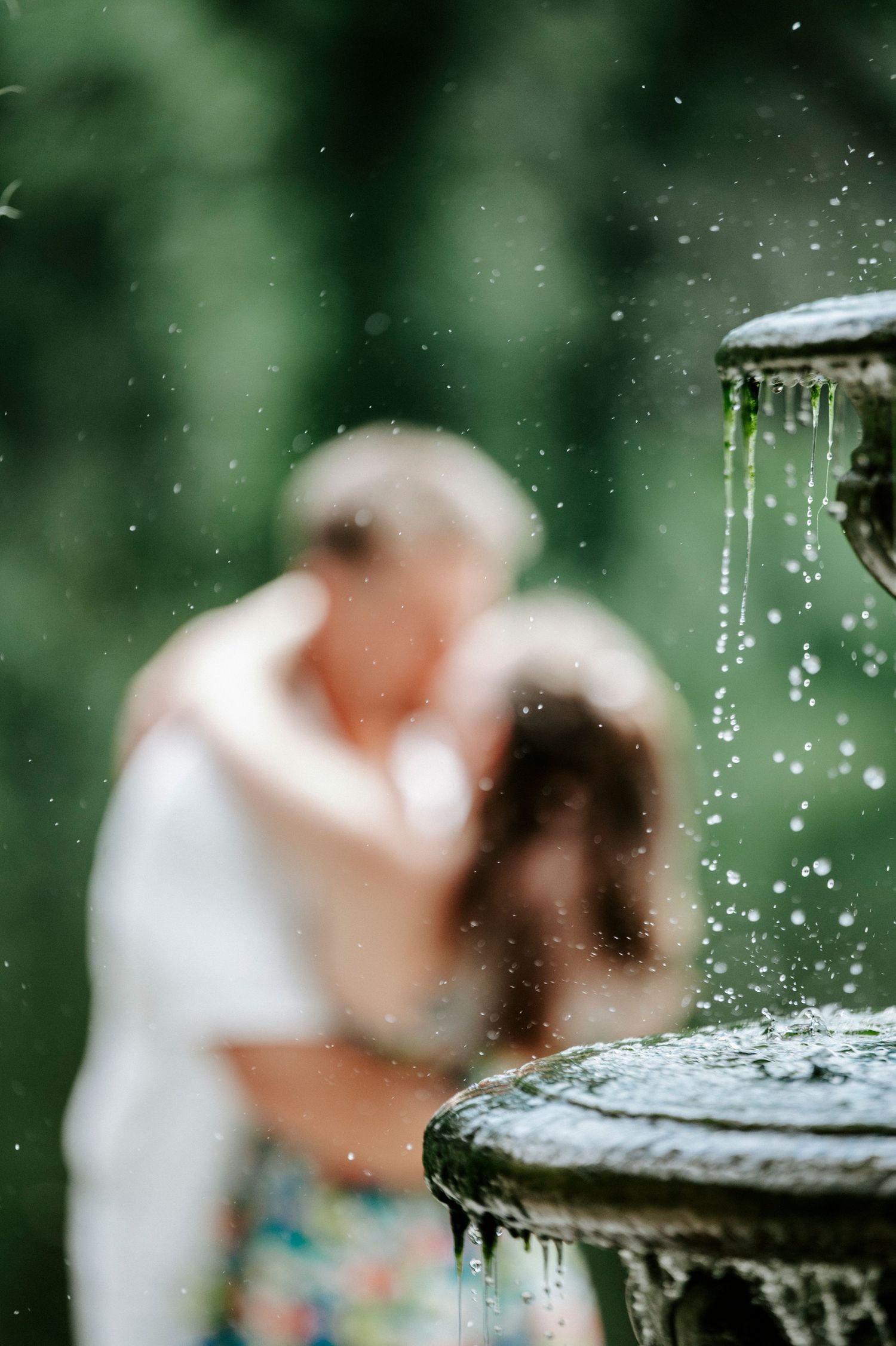 A romantic couple embraces near a fountain with bokeh water droplets in the foreground.
