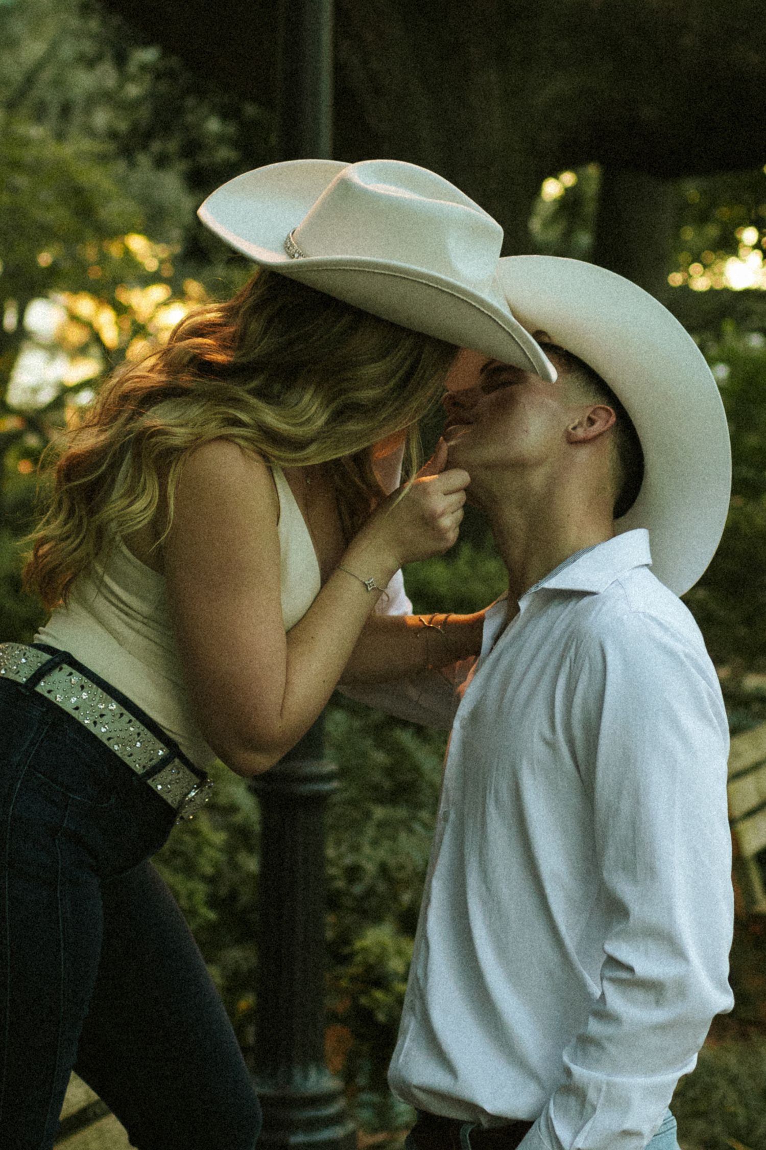 A woman standing on a bench pulls her fiance up by his chin for a kiss, they're both wearing cowboy hats
