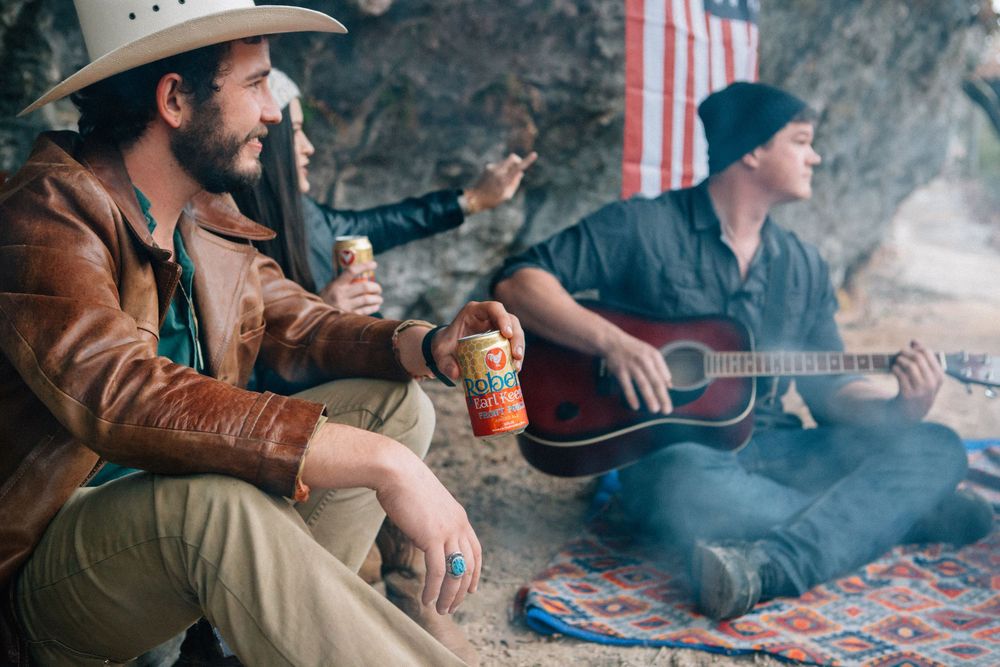 Two musicians sitting on blankets playing acoustic instruments with an American flag in the background at an outdoor gathering.