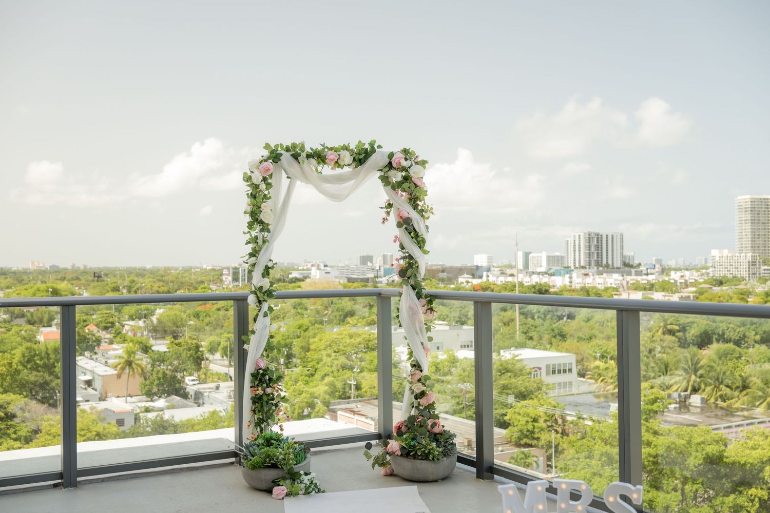A scenic urban balcony terrace decorated with potted plants and floral arrangements overlooking a city skyline on a sunny day.