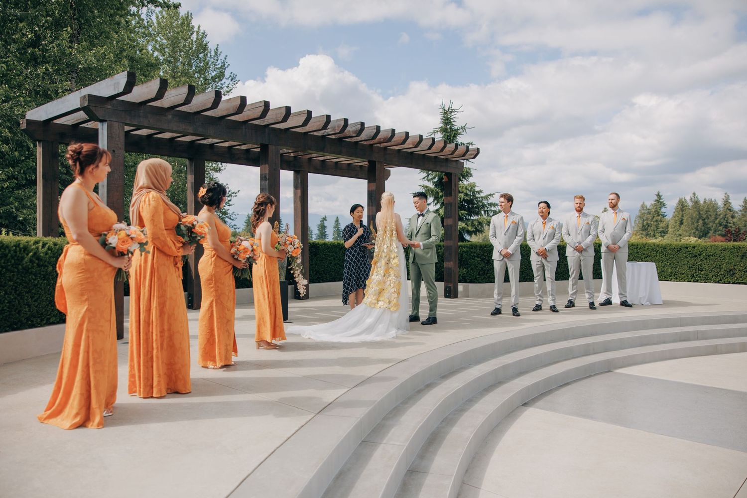 Wedding party in orange dresses poses on an elegant outdoor terrace with white chairs and pergola.