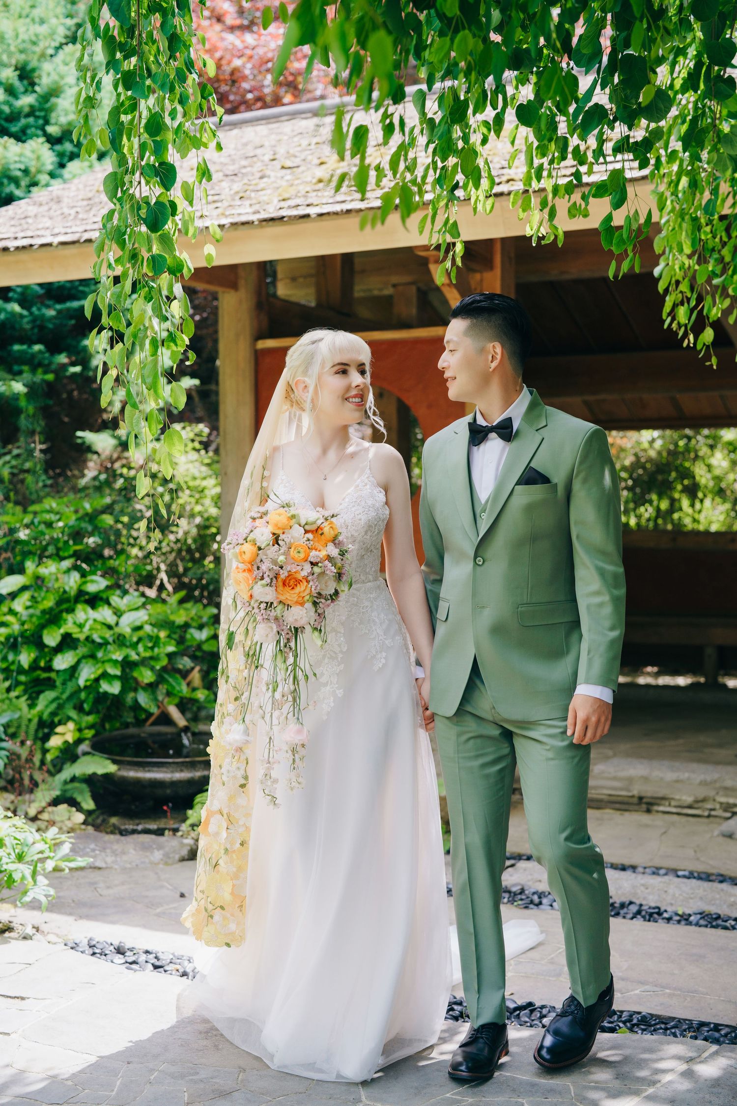 Couple in wedding attire stands together under lush green foliage during outdoor ceremony.