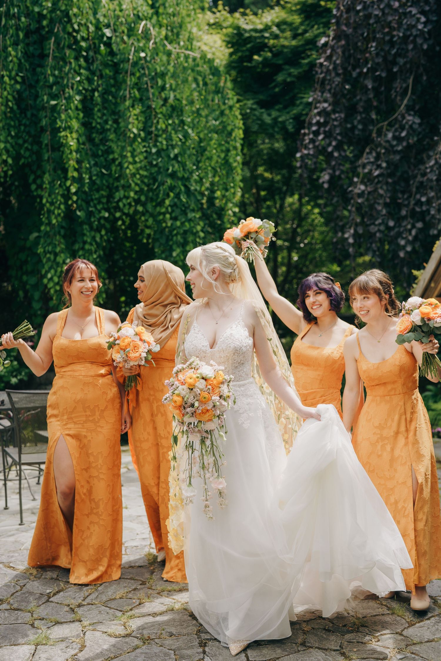 Bridesmaids in orange dresses pose playfully with bride in white gown against green garden backdrop.