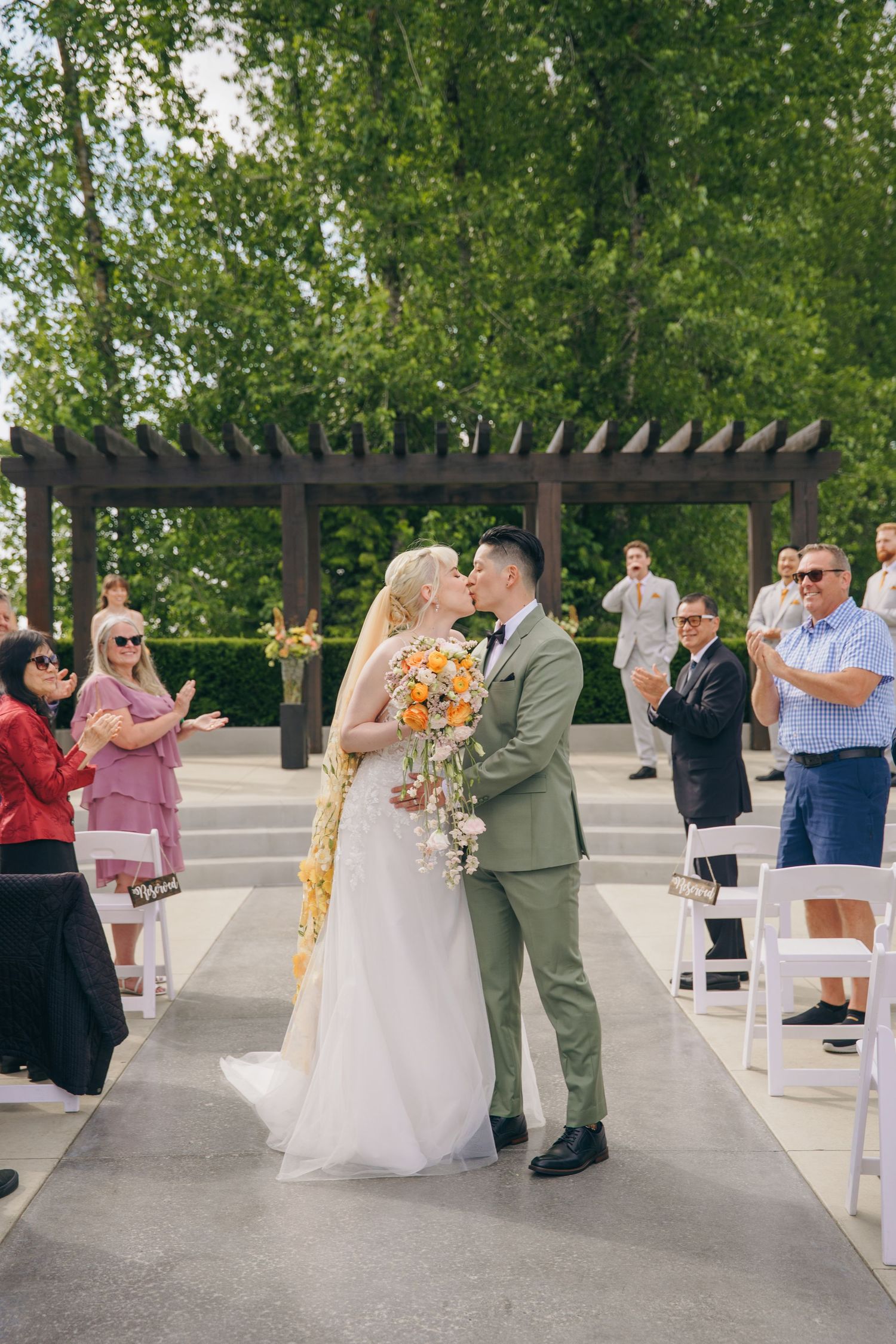 Outdoor wedding ceremony setup with white chairs and wooden pergola surrounded by trees.