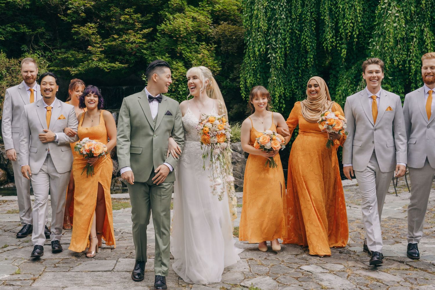 Wedding party in orange and sage green attire poses together outdoors.