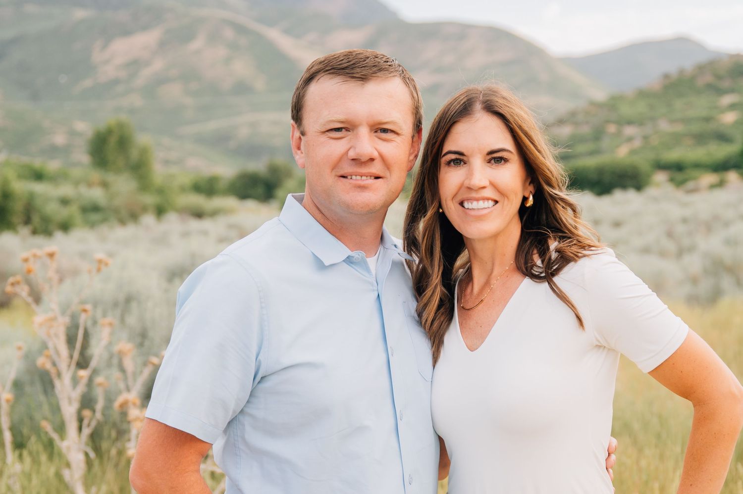 A couple poses together in an outdoor mountain setting during a photo session wearing light-colored outfits.