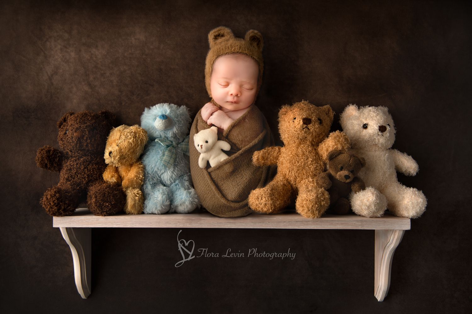 Newborn baby sleeping peacefully on neutral shelf surrounded by collection of soft teddy bears against dark background.