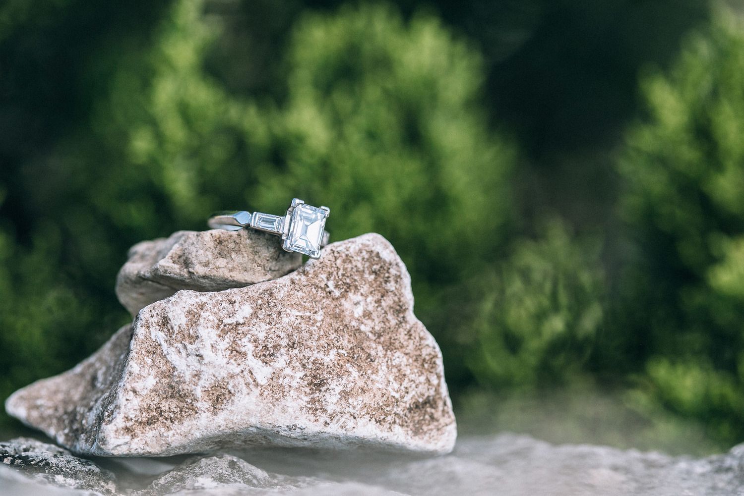 A sparkling diamond engagement ring resting on a rugged rock with lush green foliage in the background.