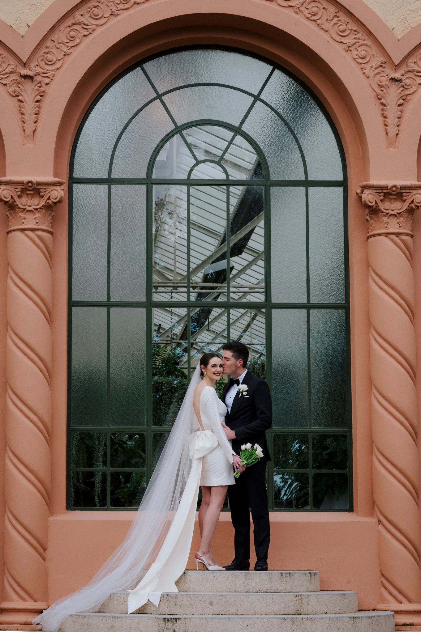 Elegant couple poses on steps in front of pink conservatory building with arched windows.