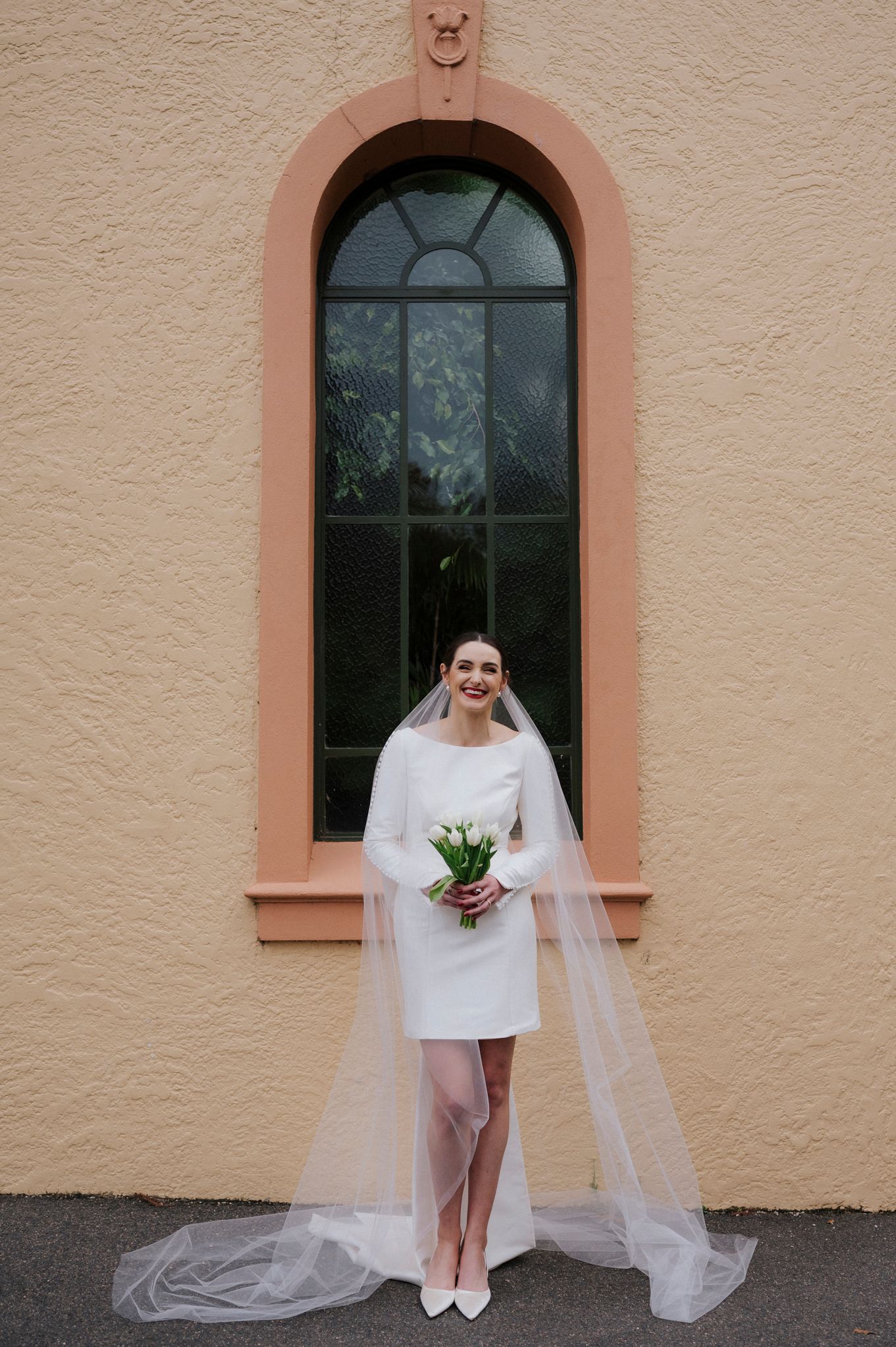 Modern bride in short dress and long veil poses against peach-colored building with arched windows.