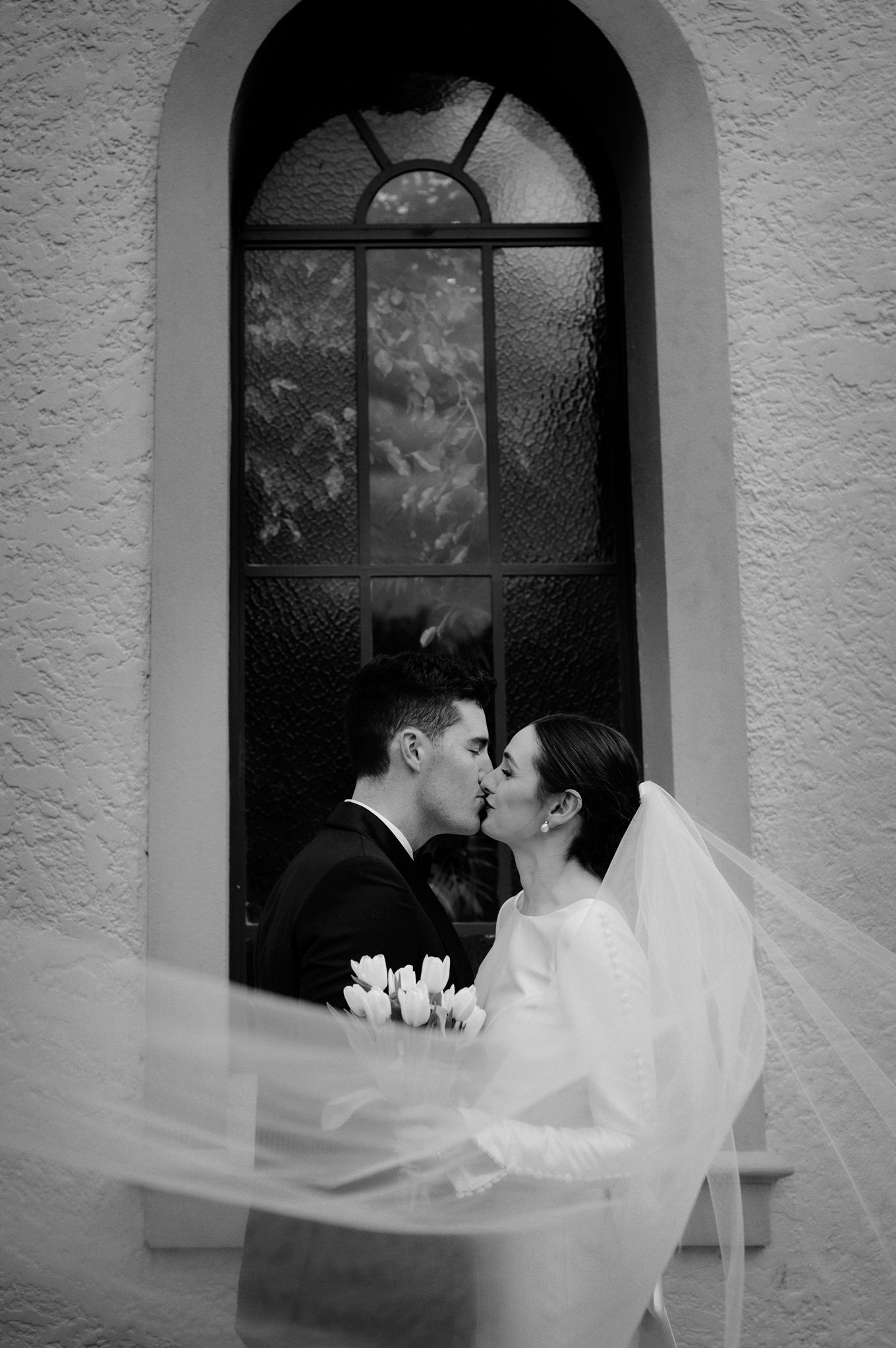 Black and white romantic moment captured through arched window of historic building.