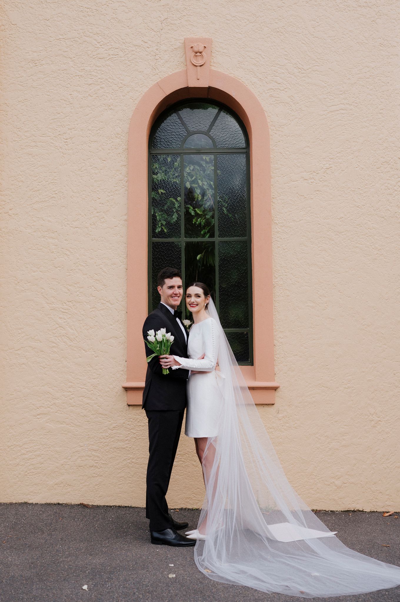 A newlywed couple poses in a pink arched doorway with a long flowing veil.