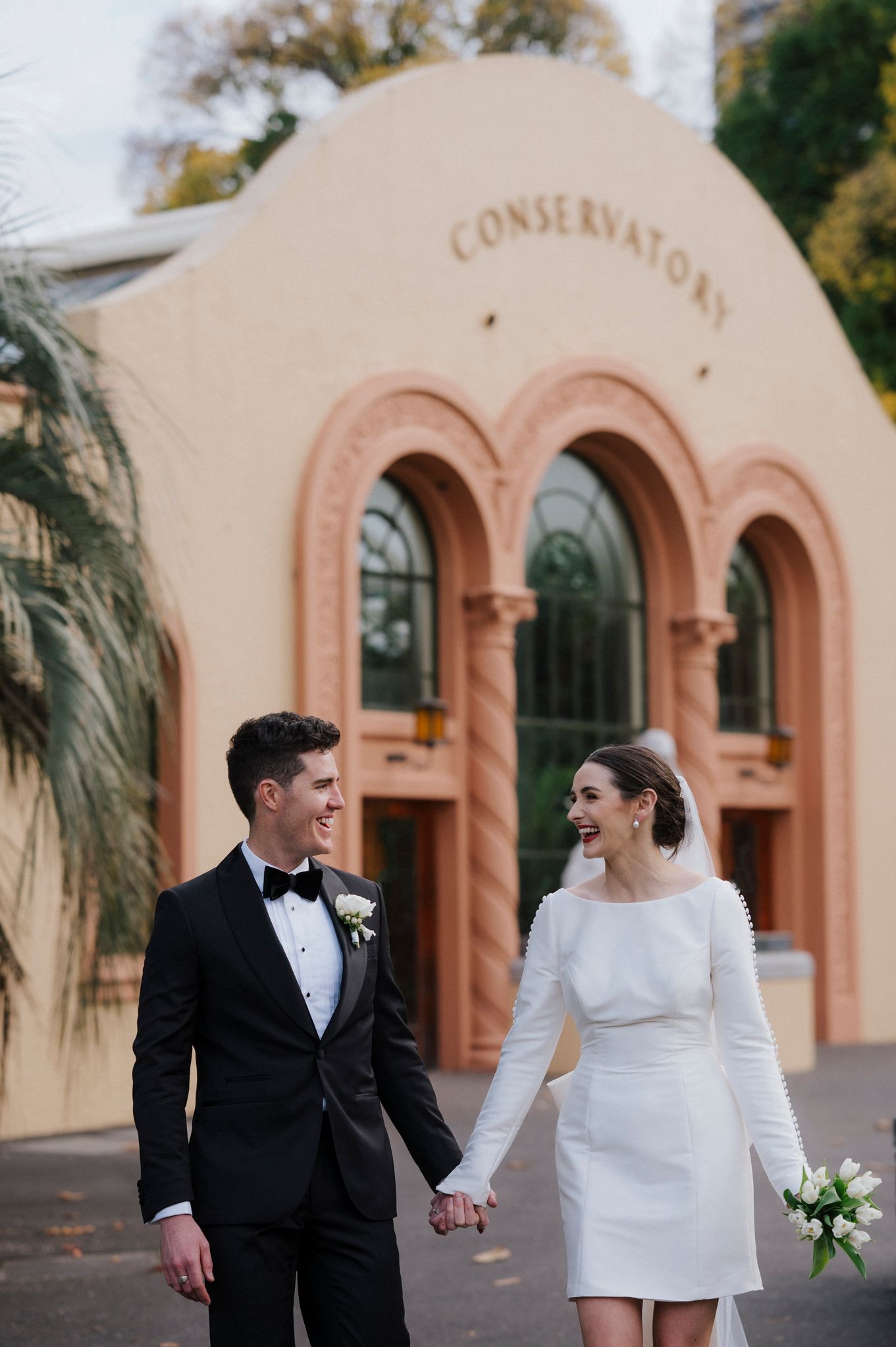 A couple in wedding attire stands smiling in front of a pink conservatory building with arched windows.